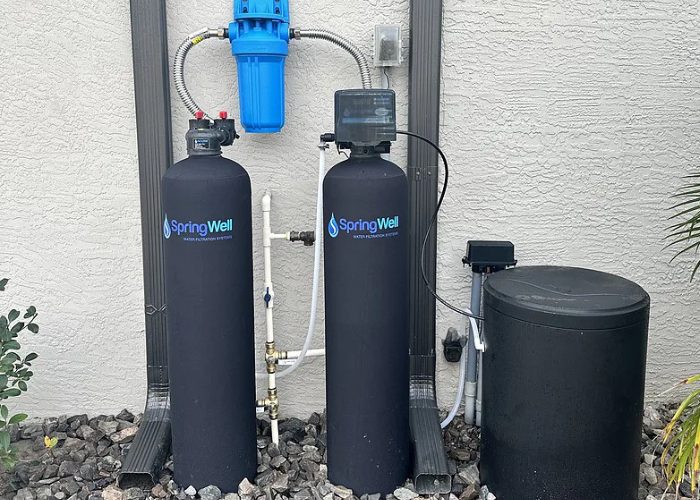 SpringWell whole house water filter and salt-based softener combo installed at an Arizona home: two black tanks with the softener's valve head, round brine tank, and blue sediment pre-filter overhead